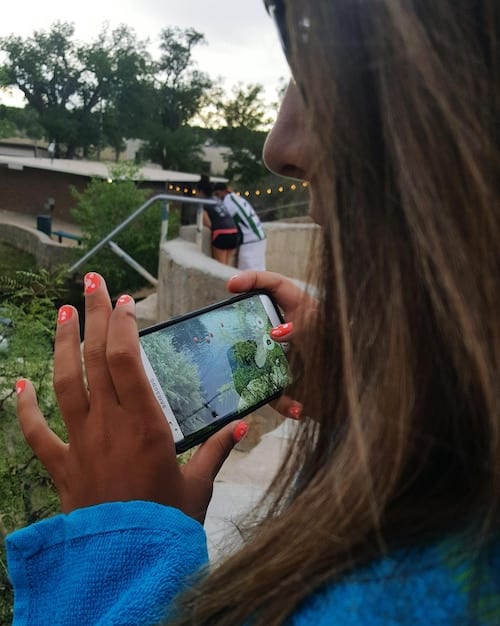 A hand holding a smartphone with a project management app displayed on the screen, showing tasks, timelines, and team communication features, set against a blurred background of a community garden.