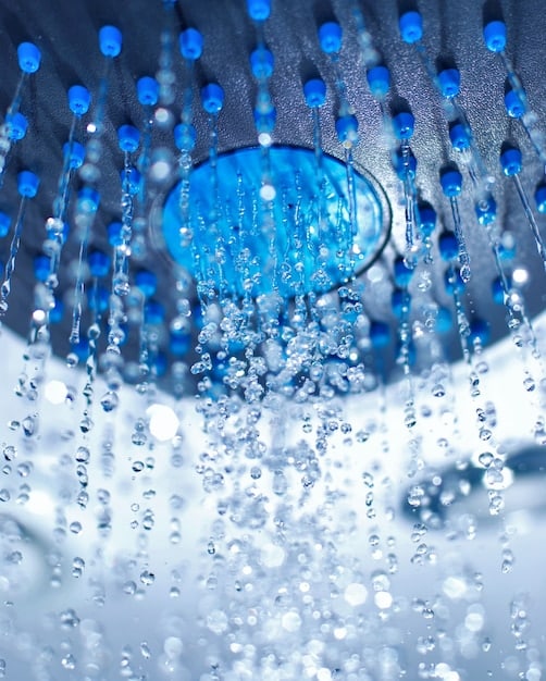 Close-up of a low-flow showerhead demonstrating water efficiency, with water droplets coming out. The background is a blurred bathroom setting.