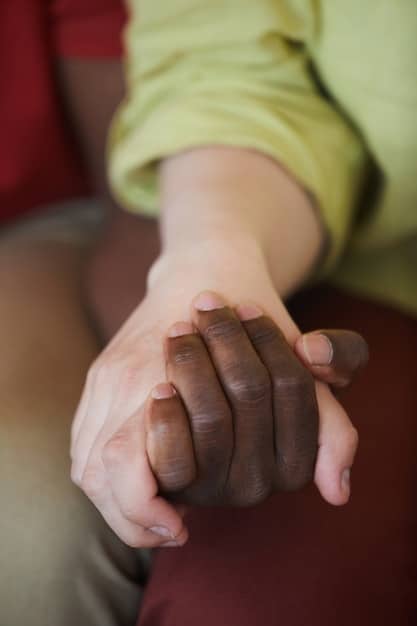 A close-up shot of a volunteer's hand gently holding the hand of an elderly patient in a hospital bed. The focus is on the hands, conveying a sense of comfort, empathy, and human connection. The background is blurred to keep the attention on the hands.